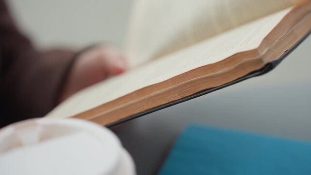 Close up shows person holding open book focusing on text filled page. white cup appears in some frames near bottom left corner. scene is softly lit with shallow depth of field emphasizing
