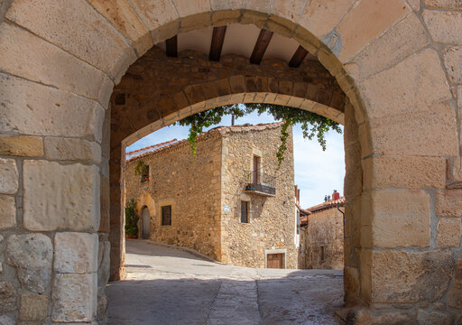 Arco de piedra que enmarca una calle tradicional de Puertomingalvo, Teruel, con casas r&uacute;sticas y encanto medieval bajo la luz c&aacute;lida del sol, s&iacute;mbolo del patrimonio y la historia espa&ntilde;ola.