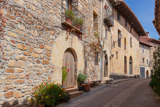 Calle empedrada con casas de piedra y balcones floridos en Puertomingalvo, Teruel, reflejo del encanto rural y la arquitectura tradicional espa&ntilde;ola bajo un cielo azul despejado