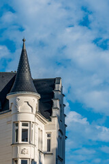 Historic European residential building architecture detail with a conical slate roof turret and decorative dormer windows against a blue cloudy sky © Quality by Simon