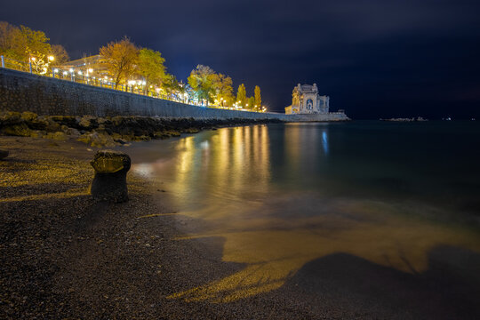 View of the Casino of Constanta illuminated at night along the Black Sea, with reflections on the water and a dark pebbled beach in the foreground Constanta, Judetul Constanta, Romania.