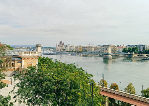View of the Szechenyi Chain Bridge spanning the Danube River with the Hungarian Parliament Building in the background under a soft sky in Budapest, Hungary.