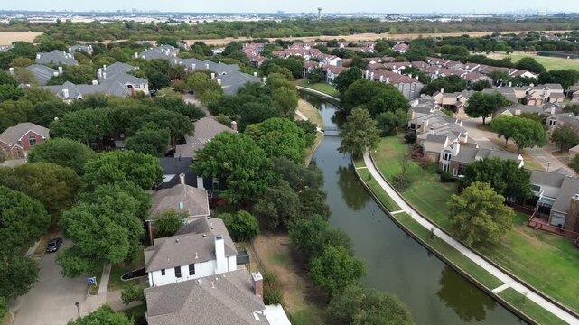 High angle perspective tracks curving canal waters running past suburban neighborhoods. Overhead altitude captures shade trees flanking residential backyards within, Valley Ranch, Irving, TX