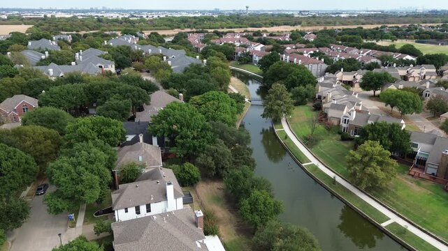 High angle perspective tracks curving canal waters running past suburban neighborhoods. Overhead altitude captures shade trees flanking residential backyards within, Valley Ranch, Irving, TX