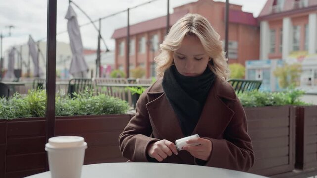 Young blonde woman sits outdoors in casual attire with brown coat and black scarf at outdoor table. Woman interacts with small objects likely related to personal grooming or beauty routine