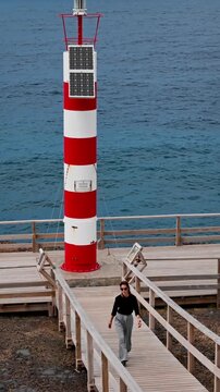 Female traveler visiting coastal lighthouse in Azores, Portugal. Vertical footage of woman walking on wooden boardwalk with striped lighthouse and Atlantic Ocean
