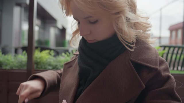 Shows lady with blonde hair removing lint from brown fabric on back of air blower while adjusting her dark scarf. Woman wears thick textured winter outfit in soft natural light