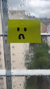Sad face sticky note on rainy window vertical stuck indoors