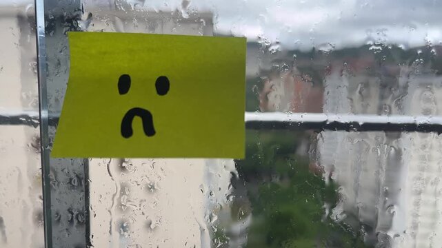 Sad face sticky note on rainy window stuck indoors