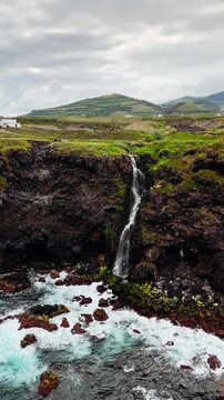 Waterfall dropping from coastal cliff into ocean with volcanic stones and moss, aerial view Azores island coastline