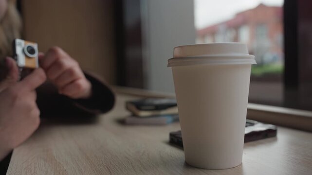 Woman sits by window holding compact camera. white takeout coffee cup stands on wooden table beside her with stacked notebooks or devices in soft focus. scene is calm indoors