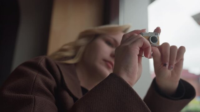 Lady with blonde hair wearing brown coat carefully frames and captures images using vintage style miniature camera inside room. soft lighting highlights her hands and small device
