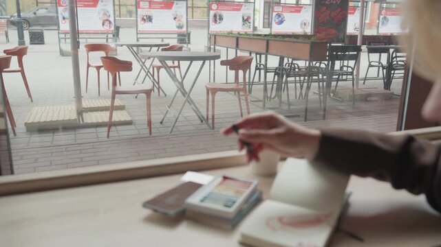 Hand reaches for art supplies on table then draws in notebook. scene is set indoors with soft lighting featuring empty cafe furniture visible through window