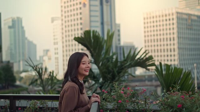 Happy young Asian woman walking onto urban balcony and enjoying city view, smiling female relaxing among greenery with modern buildings in background, outdoor