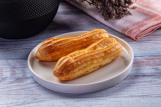Golden baked eclair pastries on white ceramic plate with textured black speaker and dried lavender on striped cloth background, dessert