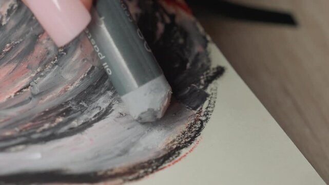 Close up shows person pink nailed hand using oil pastel labeled 09 ceram to apply soft gray color over textured paper with swirling patterns and red accents. scene focuses on interaction