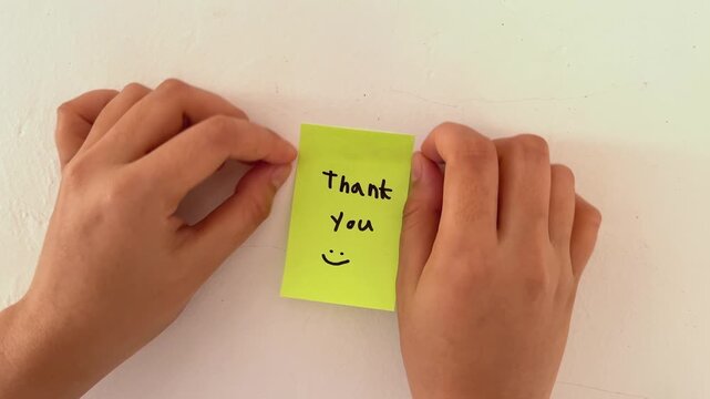 Woman hands gently place a sticky note on a clean wall. Yellow note reads "Thank You" with a cheerful smile. Simple gesture shows gratitude and warmth