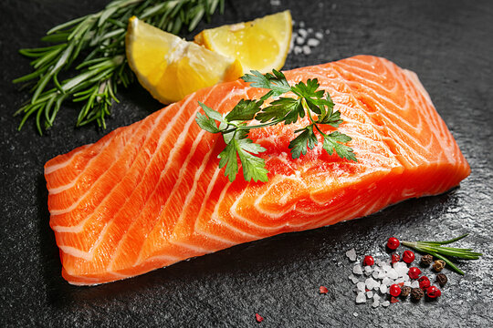 Fresh raw salmon fillet with lemon, rosemary, parsley, salt and pepper on a black slate surface