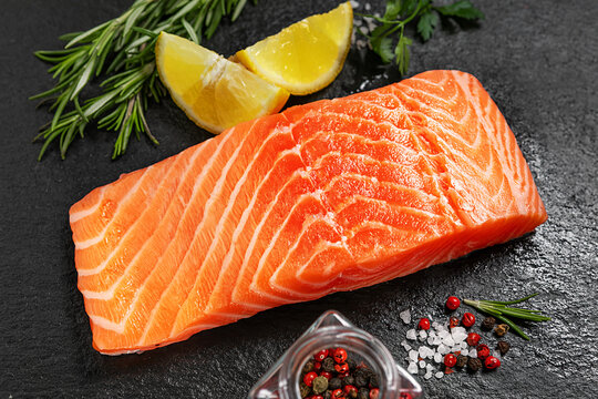 Fresh raw salmon fillet with lemon, rosemary, parsley, salt and pepper on a black slate surface