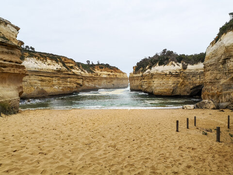 Sandy beach framed by towering limestone cliffs opening to the sea in a narrow gorge under pale sky, Loch Ard Gorge, Victoria