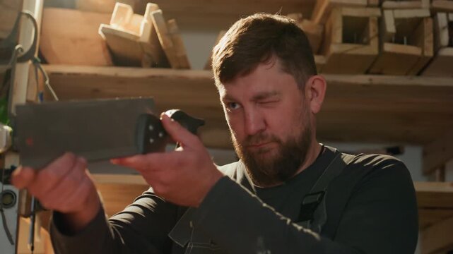 Man wearing overalls uses handheld electronic device to measure wooden materials. Workshop shelves filled with lumber and tools visible warm lighting creates focused shadows
