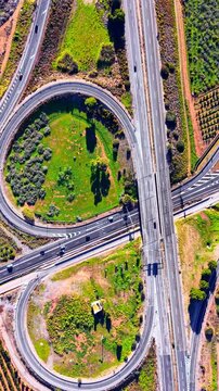 Highway cloverleaf ramps and overpass with green infield, Spain. Looping slip road around a grassy infield with shrubs, a multilane highway bridge, road markings and a few cars. Vertical video.