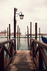 A weathered wooden walkway extends toward gondolas and mooring posts on the Venetian Lagoon. © Dillon T.