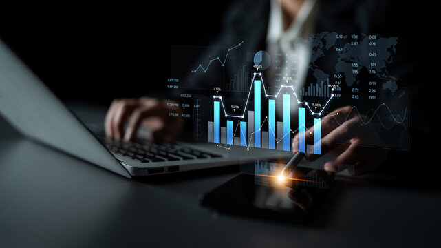 A business professional engages with financial data, utilizing a laptop and smartphone to analyze charts and graphs, highlighting modern technology in a dark office setting. Scalp