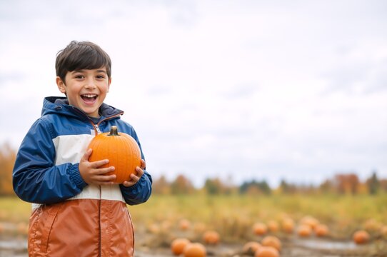 Happy young boy holding an orange pumpkin in a field. Smiling child in a rain jacket at an autumn pumpkin patch