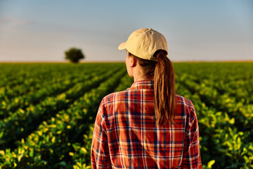 Fototapeta premium Woman farmer wearing a cap and plaid shirt standing in a lush green soybean field, overseeing healthy agriculture and future harvest