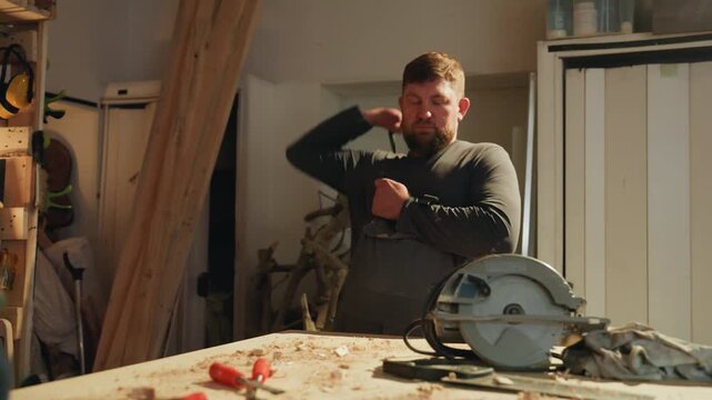 Man stands at workbench filled with wood shavings. Man adjusts his apron straps while various hand tools like pliers lie on table surrounded by wooden shelves and workshop equipment