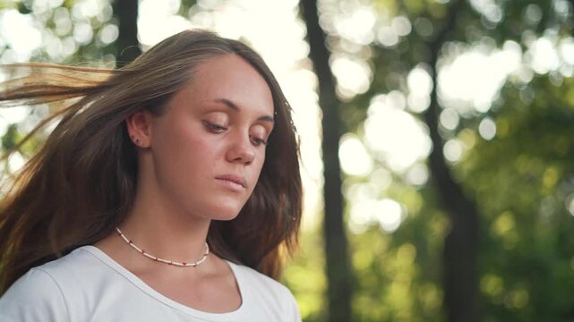 Young woman meditating peacefully in park during golden hour. Female relaxes outdoors with eyes closed. Girl enjoys calm meditation in nature. Woman practices mindfulness under trees with sunlight.
