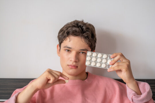 Young adult holding single pill and blister of medication, habit repeated at first symptom, story told without any single spoken word. Wide shot. Concept of medication taken at home without doctor