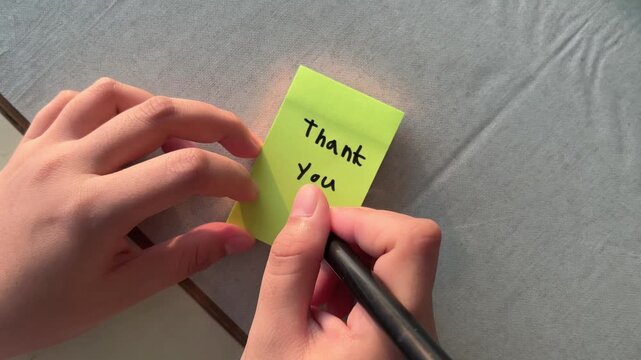Woman hands write "thank you" on yellow sticky note. Fingers press down note carefully.