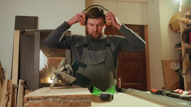 Man wearing overalls picks up yellow ear protection muffs from workbench preparing to use circular saw on wooden board. Workshop background includes various woodworking materials like planks
