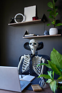 Skeleton figure wearing glasses and tie sitting at desk with laptop and notebook