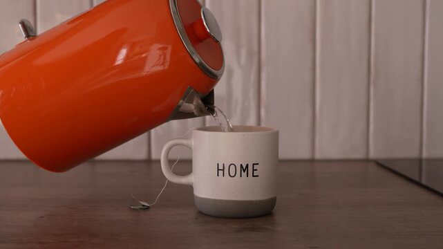 Young woman pours hot water from an orange kettle into a mug with a tea bag, brewing warm tea at home in a cozy kitchen