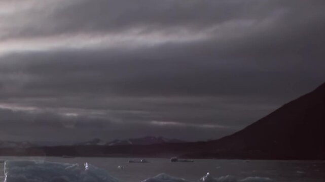 Rugged mountains rise above cold water scattered with floating ice. The frame preserves overcast light and a dark bay as supporting detail, adding natural texture and depth to the view.