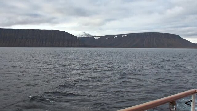 Polar Ocean Seascape Moving Under Cold Northern Sky Across Open Water. Snow-covered mountains remain visible beyond the cold open water and the distant polar horizon.