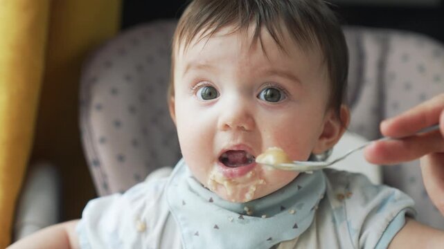 Adorable baby eating porridge with spoon in highchair, food all over face and chin. Parent feeds infant during mealtime as child learns to eat solid foods. Wearing blue bib for messy eating.
