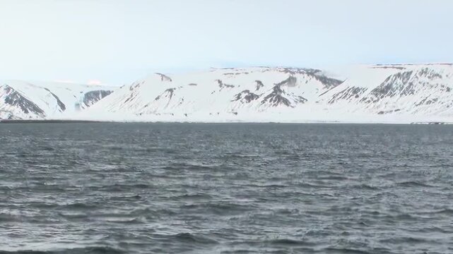 Cold Arctic Waters Glowing Beneath Wide Polar Sky Under Changing Light. Snow-covered mountains remain visible beyond the cold open water and the distant polar horizon.