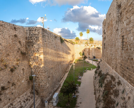 Historic stone fortress walls surround a peaceful pathway in Acre, Israel, with palm trees, dramatic clouds, and blue sky.