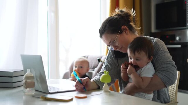 Mother working from home with toddler and baby nearby. Woman multitasks writing notes with laptop while parenting. Mom balances remote work with childcare. Parent manages kids and work.
