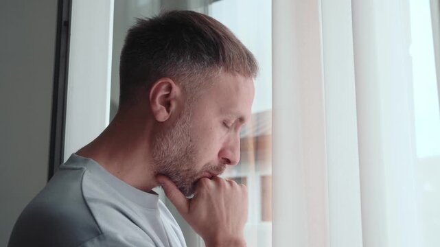 Anxious man stands by window with hand on chin, looking worried and overwhelmed while thinking through stress, uncertainty and difficult decision at home.