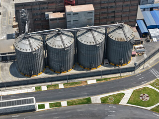 Four vertical metal grain silos beside a large industrial building and an avenue with green areas, aerial view © WILL PHOTOGRAPHY