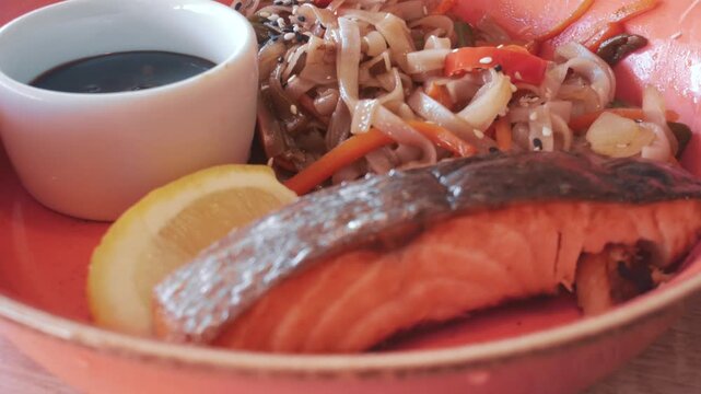 Grilled salmon served with noodles, vegetables, sesame, lemon and savory sauce as balanced seafood meal for lunch or dinner.