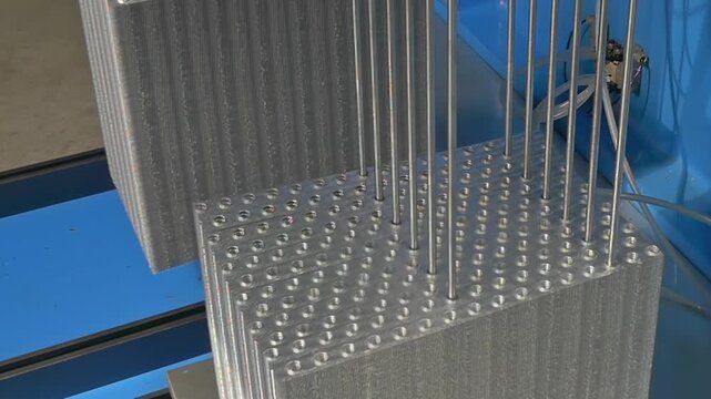Slender steel rods descend into geometric holes in a metallic block, showcasing mechanical synchronization and high-tech engineering excellence during a daytime assembly process.