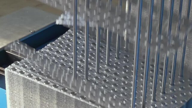 Shiny steel plates move beneath vertical rods within a heavy manufacturing facility during the bright afternoon to create precision patterns for construction components and structural supports