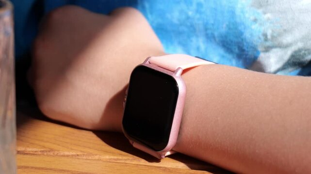 Pink square smartwatch with blank black screen on a wrist resting on a wooden table, pale pink silicone band, blue denim background, bright sunlight, closeup, no face, technology lifestyle
