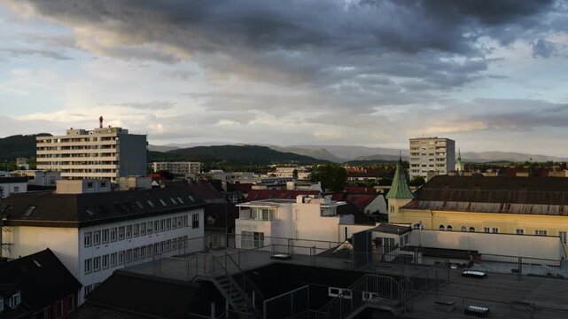 Clouds shift in the sky as light changes over buildings in the city of Klagenfurt during sunset.